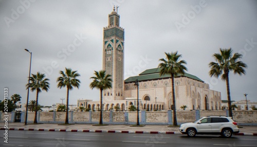 Foto The Hassan II Mosque in Casablanca, Morocco, featuring its iconic minaret and traditional architecture surrounded by palm trees under a cloudy sky