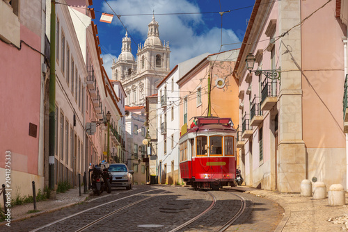 Traditional red tram travels along a narrow street in Lisbon with the National Pantheon visible in the background.