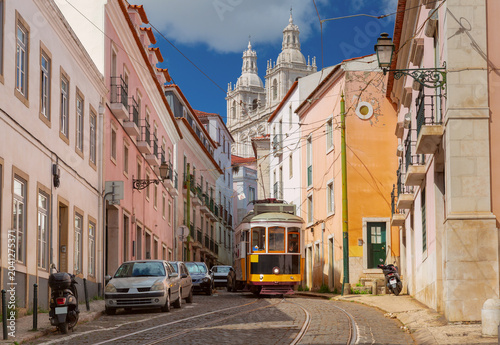 Traditional yellow tram travels along a narrow street in Lisbon with the National Pantheon visible in the background.