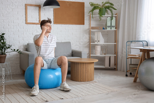 Young man talking by mobile phone on fitball at home