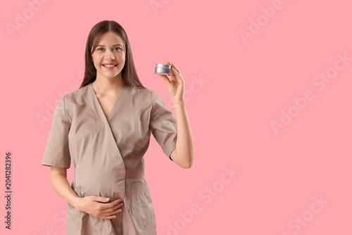Young pregnant cosmetologist with jar of cream on pink background