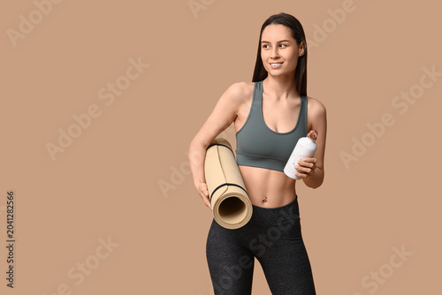 Young sporty woman with water bottle and yoga mat on beige background