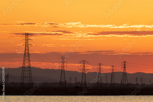 a dramatic sunset landscape featuring a line of high-voltage electrical transmission towers silhouetted against a brilliant orange and gold sky. Sunnyvale Bay Trail, Santa Clara County, California.