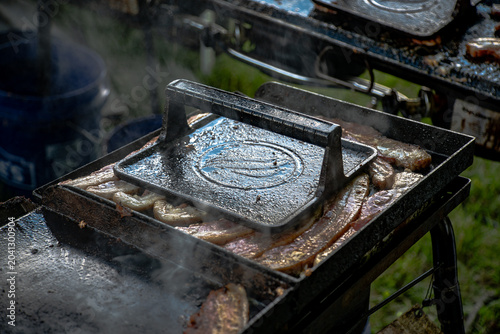Obraz na płótnie Bacon Cooking Under Cast Iron Press on Outdoor Flat Top Grill