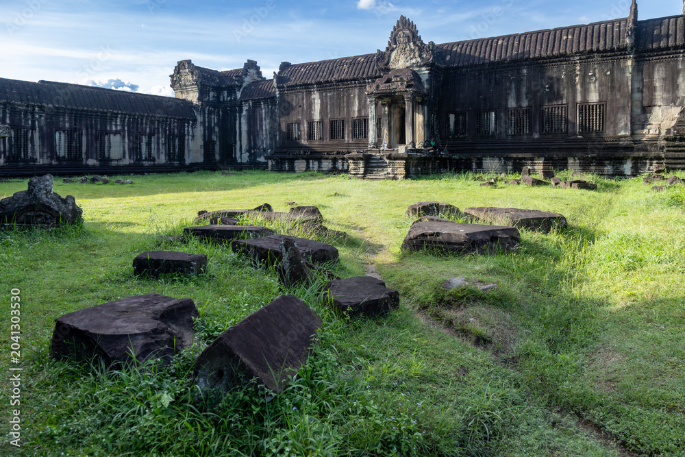 Fototapeta premium Angkor Wat, Siem Reap, Cambodia. Ancient stone ruins lie scattered on the grass in front of the temple complex, bathed in soft sunlight under a blue sky.