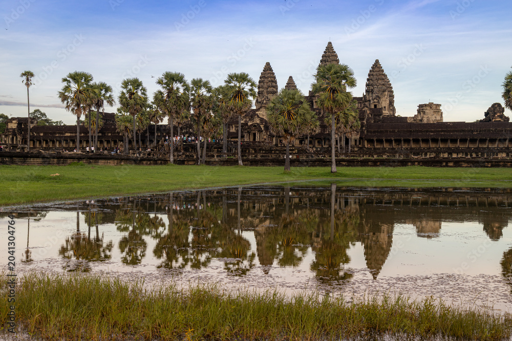 Fototapeta premium Angkor Wat, Siem Reap, Cambodia. Tourists admire the iconic temple complex at sunrise, reflected in the surrounding moat. The ancient Khmer architecture stands majestically against a soft sky.