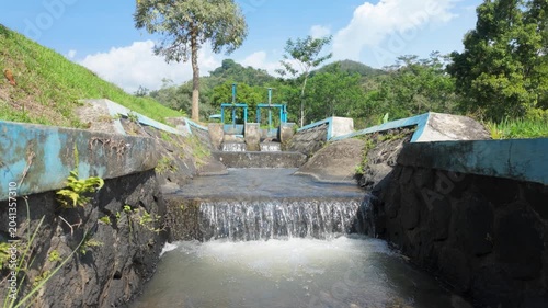Water flowing over a stone pathway surrounded by greenery in a sunny landscape