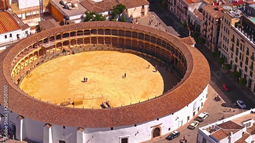 Drone footage panning sideways and tilting down into Ronda's famous bullring, revealing the perfect golden sand floor, tiered arched seating, rooftop pool, and visitors dotted inside.