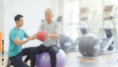 A trainer assists a senior individual in a gym, focusing on strength training with a medicine ball while seated on a stability ball in a bright environment.