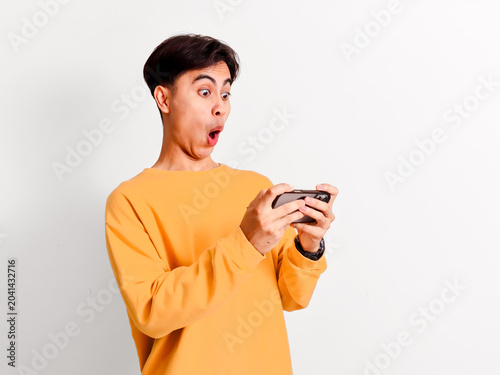 Excited young Asian man playing mobile game on smartphone with funny open mouth expression on white background