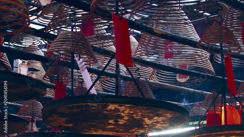 Arc left showing the ambient motion of smoke rising from layered spiral incense coils hanging from the ceiling with red tags decoration for Lunar New Year in Man Mo Temple. Sheung Wan, Hong Kong