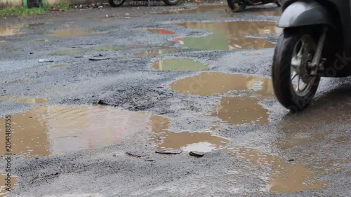 A damaged neighborhood street covered with rainwater puddles and deep potholes as riders on scooters maneuver carefully through the uneven and hazardous surface.