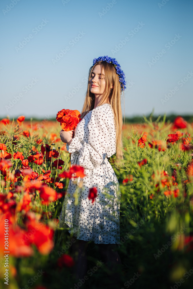 Obraz premium Blonde slavic female posing in white dress, blue flower crown, holding red poppy bouquet amid sunlit summer meadow