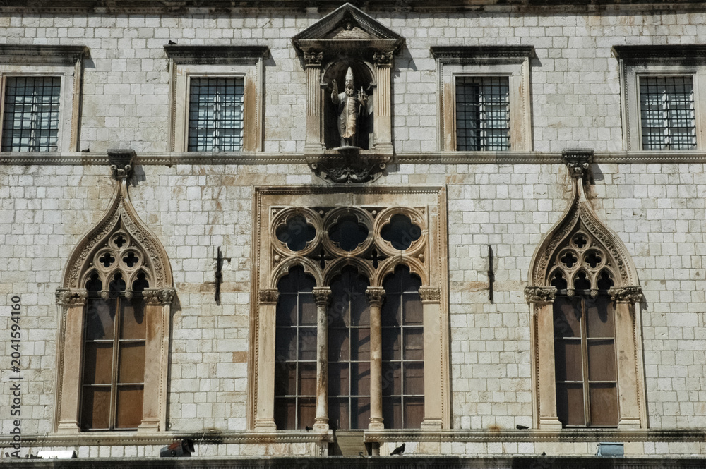 Obraz premium Facade of Sponza Palace in Dubrovnik showing historic Gothic and Renaissance windows with a statue of Saint Blaise on the stone wall