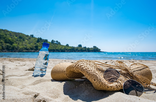 A straw hat, sunglasses, and a water bottle are placed on sandy beach. The sea is visible in the background under a clear blue sky. It represents summer vacation.