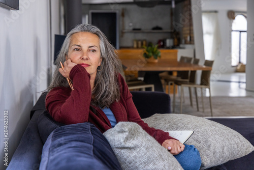 Senior woman sitting on blue sofa at home in maroon cardigan holding pencil, notebook, copy space