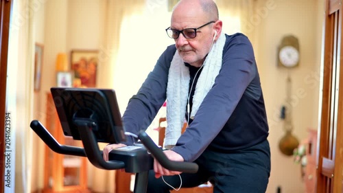 Senior Man Exercising On Stationary Bike At Home Wearing Headphones And Towel