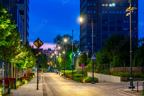 Modern city street in Lodz at dusk with cobblestone pavement, illuminated by warm street lamps, surrounded by contemporary office buildings, rows of young trees, benches, and road signs under a dark b