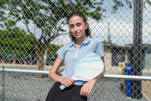 Happy young woman holding pickleball paddle racket standing near wire mesh fence. Smiling female athlete portrait posing on outdoor padel tennis court arena
