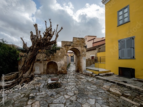 Wide view of a Plaka courtyard in Athens, featuring a stone arch, cobbled ground, and a vivid yellow ocher house beside historic masonry.