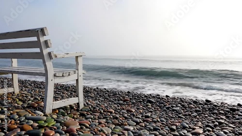 Tranquil view of white bench on a pebble beach overlooking the misty ocean with gentle waves under a bright sky