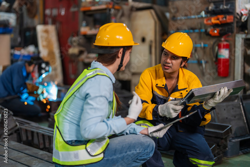 Two workers wear yellow hardhats and safety vests. They sit on a wooden bench in a workshop. One holds a metal plate while the other points to notes
