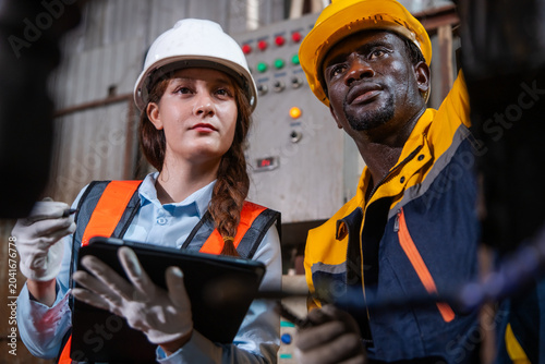 Female worker wears white hard hat and orange safety vest. She holds a tablet computer. Male worker in yellow hard hat and blue work shirt looks on. Industrial control panel with green