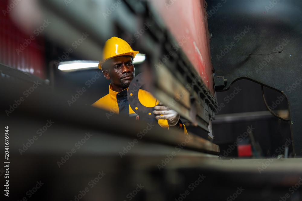 Fototapeta premium Worker wears a yellow hard hat and work jacket. He stands beside a large train. His hands rest on a metal structure. The background features blurred workshop lights