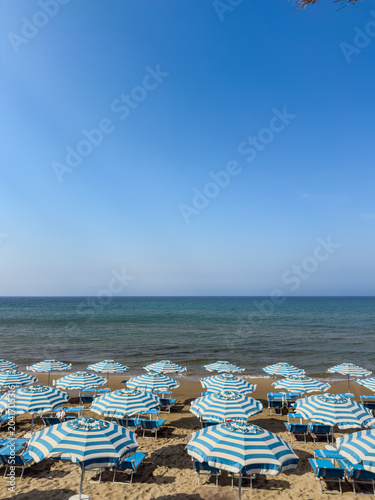 Rows of striped beach umbrellas on a sandy beach by the sea. A high-angle vertical shot of a beach scene featuring many blue and white striped umbrellas and blue lounge chairs arranged on the sand.