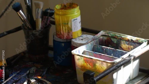 Messy artist studio table with acrylic paint brushes jars tubes and palette showing creative working process in natural light