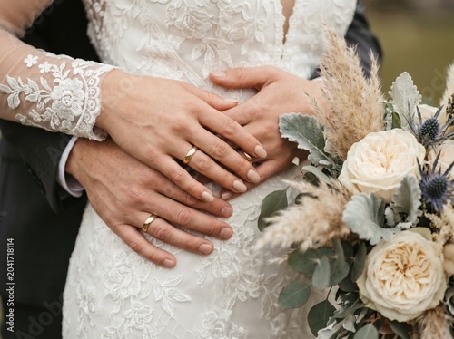 Close-up of couple holding hands on bride’s lace gown, showing wedding rings and bouquet with pale roses, eucalyptus leaves, and dried grasses