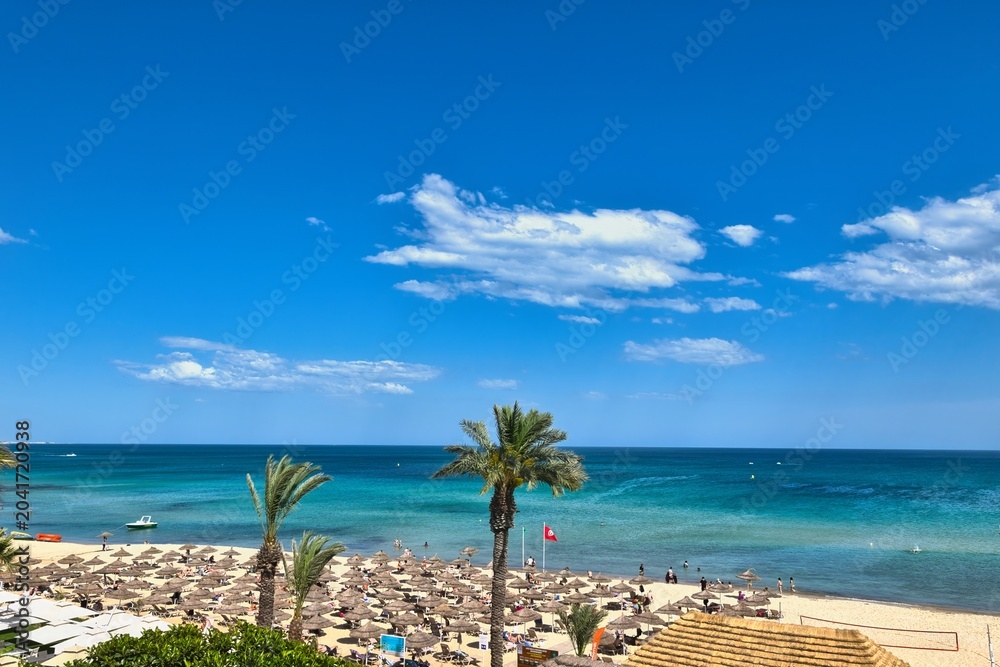 Fototapeta premium Hammamet, Tunisia - MAY 16, 2025: Beach of Hammamet, the first tourist destination in Tunisia. Located on the Gulf of Hammamet. Beach and boats on shallow water, view on Mediterranean sea. Crystal-cle