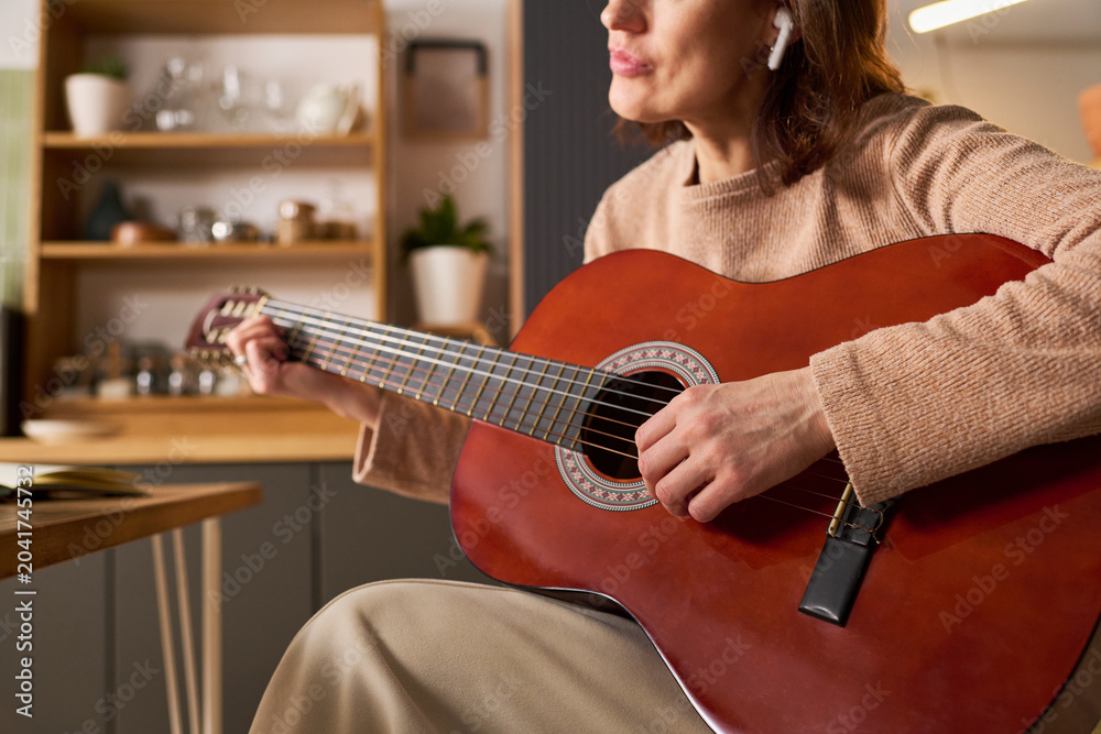 Fototapeta premium Middle aged Caucasian woman playing acoustic guitar at home while learning music online through wireless earbuds, representing remote skill building. Ideal for e learning, hobby courses