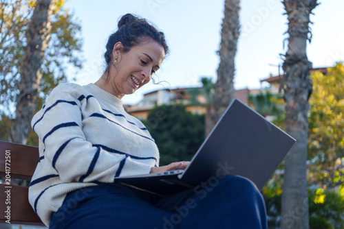 Woman smiling while using laptop outdoors, enjoying remote work and connecting digitally in a park environment