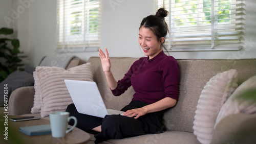 Asian woman professional remote worker doctor video call consultation using laptop computer waving greeting patient telehealth appointment home office sofa