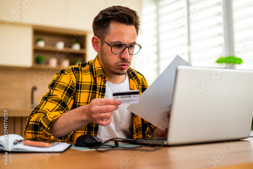 Anxious young man holds a credit card while working remotely on his laptop, appearing worried and stressed.
