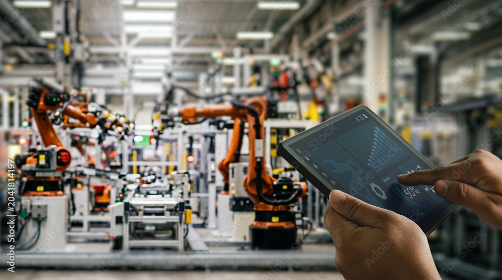 Fototapeta premium Hands holding tablet with industrial automation dashboard inside smart factory with robotic arms, supporting manufacturing monitoring, machine control, production analysis, and industrial workflow.