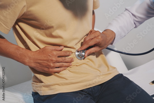 Photography A close-up shot of a doctor examining a patient's stomach area with a stethoscope to listen for bowel sounds or abdominal issues, while the patient rests his hand on his stomach