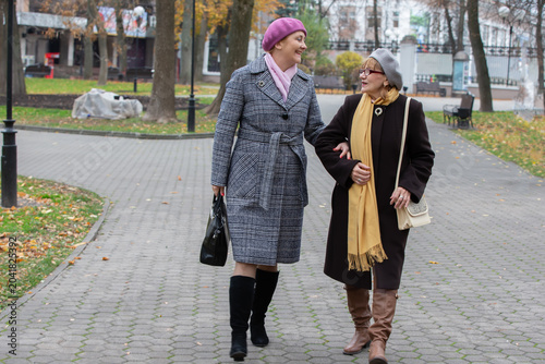 Two women walk together in a park during autumn wearing warm coats and hats