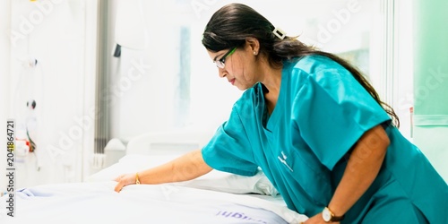 Foto Nurse in teal scrubs tending to a hospital bed in a bright room.
