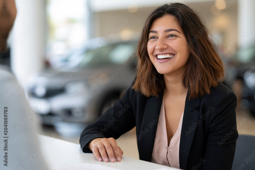 Obraz premium Warm smile brightens a woman's face. She sits at a showroom counter, attentive and pleased. Car gleams softly in the background. The setting suggests a positive, professional interaction