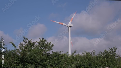 Wind turbine blades turning among green trees under a blue sky