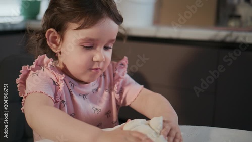 Young girl plays with dough at kitchen table. She wears pink ruffled shirt, focused and curious. Soft lighting highlights her concentration. Ideal for family, child development