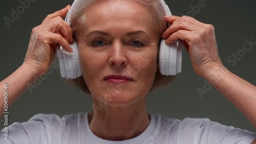 Portrait: happy female pensioner puts headsets on head. Face of senior old woman with gray hair elderly grandmother. Listening to music or podcast with white headphones. Wrinkles on face, good mood