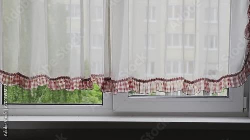 Light transparent curtain with red plaid ruffles moving gently in the breeze. A view from an apartment window showing urban scenery