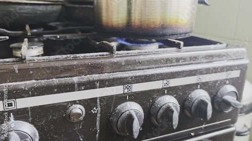 Extremely dirty and greasy old gas stove with a pot cooking on a lit burner. Unsanitary kitchen conditions representing neglect