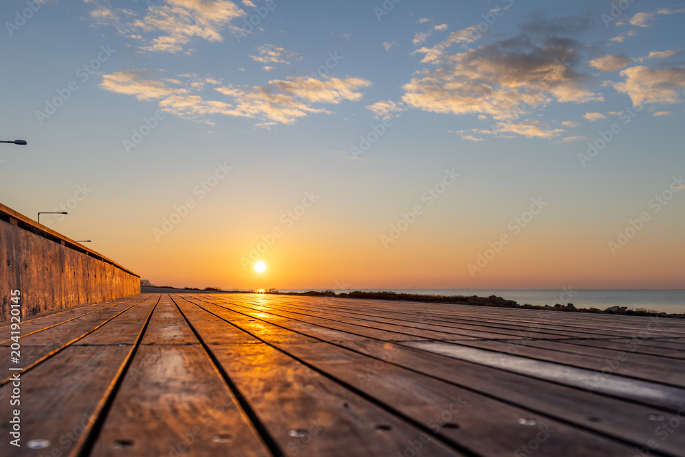 Fototapeta premium Golden morning sunrise over calm sea from low angle perspective on wooden boardwalk planks in off-season. Beautiful coastal landscape reflection. Ayia Napa, Cyprus.