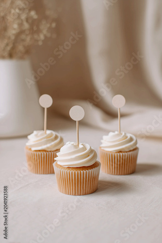 Three vanilla cupcakes with blank round wooden toppers mockup in a minimalist aesthetic neutral background for wedding or birthday party dessert presentation. AI generated