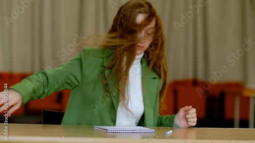 Young woman in green suit angrily tearing assignment paper at classroom table, throwing away failed attempt, theme frustration burnout and perfectionism in student life.
