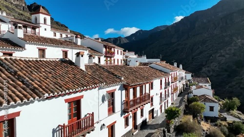 Drone view of a charming white village with red trim nestled in a stunning mountain range on a sunny day